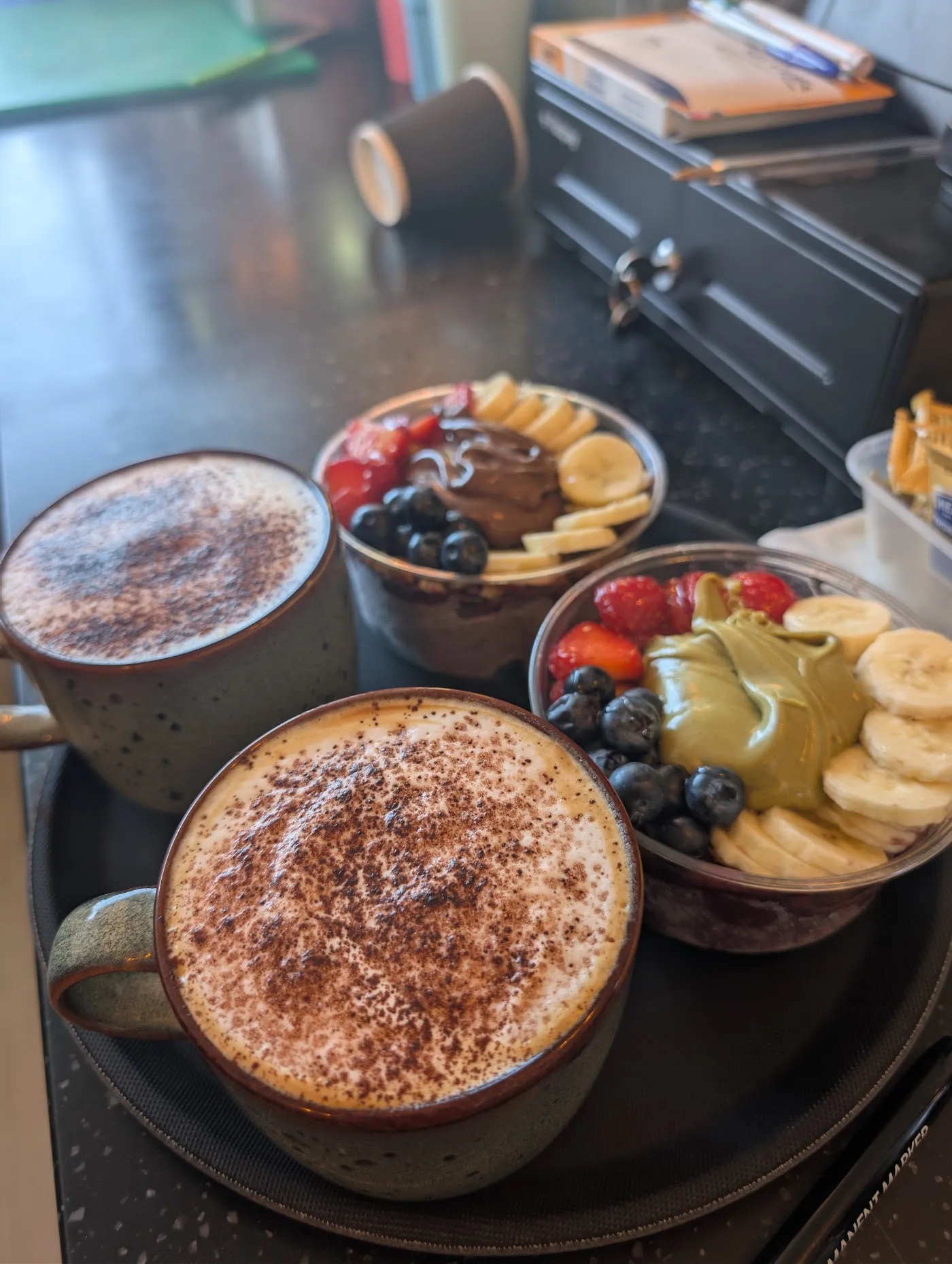 Two açai bowls topped with banana, strawberries, blueberries and nut butter, beside two cappuccinos dusted with chocolate