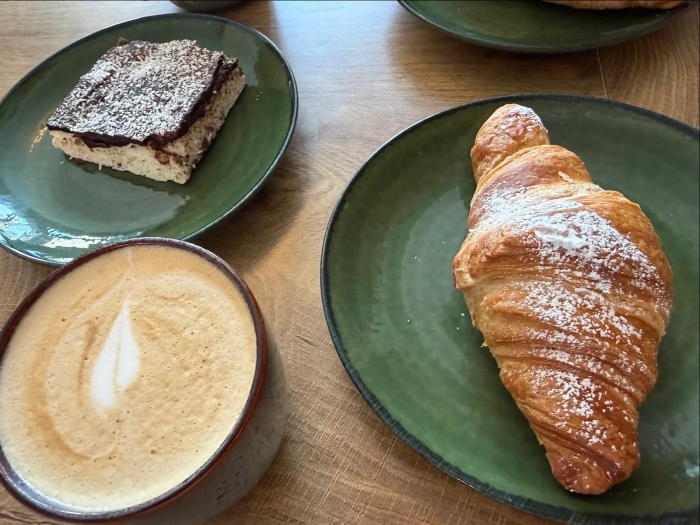 A sugar-dusted croissant and a chocolate slice on green plates beside a latte with leaf latte art, on an oak table