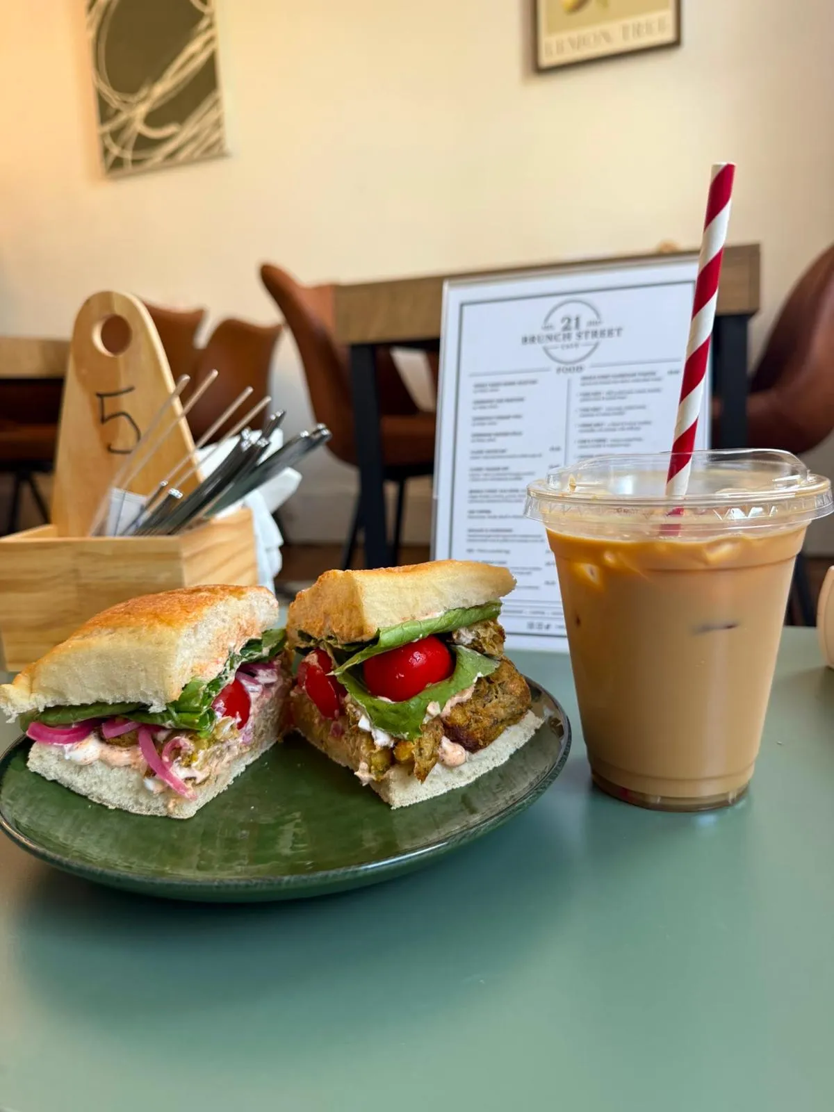 Two filled focaccia sandwiches on a green plate and an iced latte, on the sage green counter