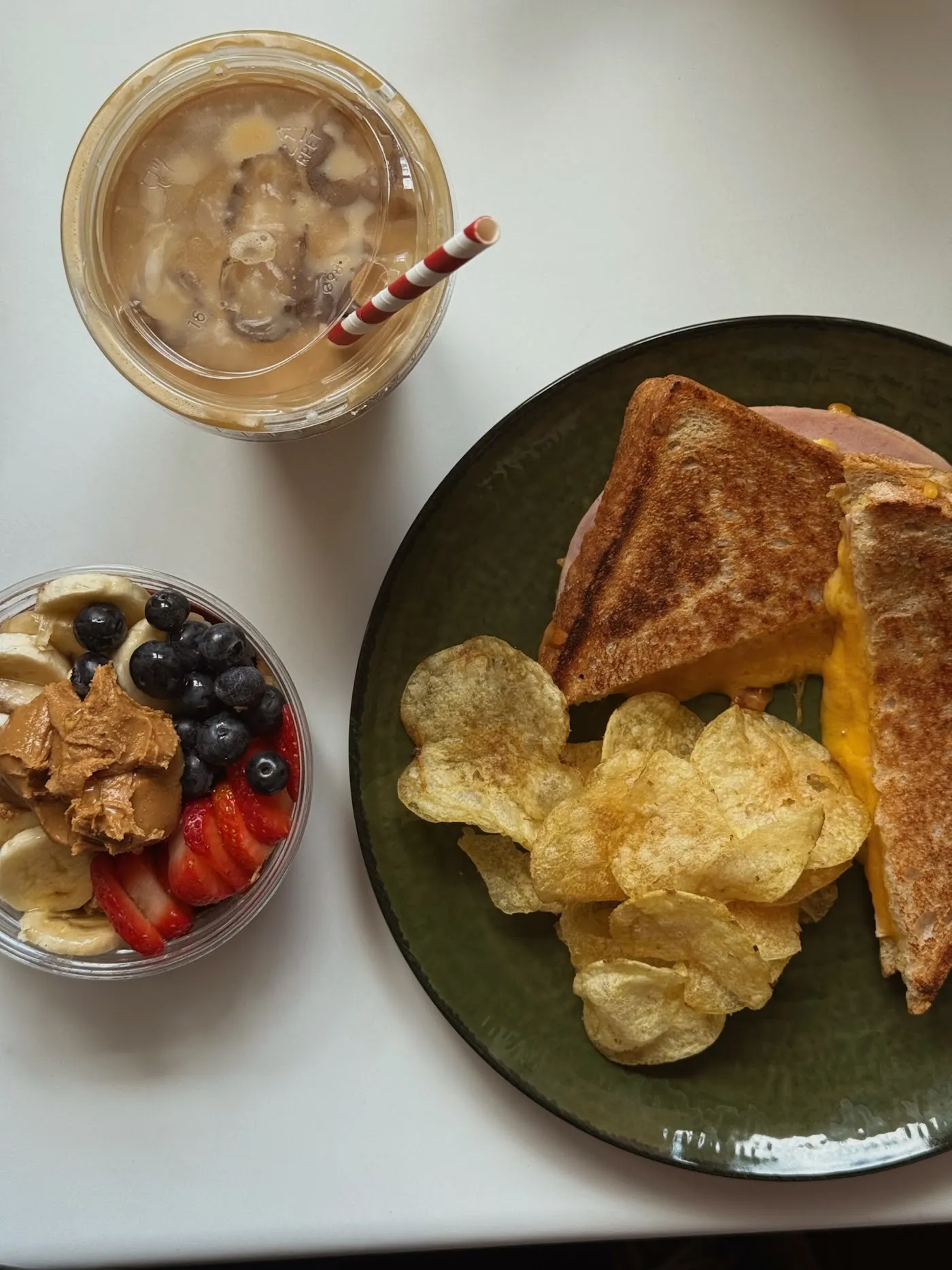A golden cheese toastie with ready salted crisps on a green plate, an açai bowl and an iced latte with a striped straw